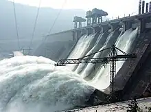 Image of a large water retaining structure that spills water like a waterfall, some power lines and a construction crane