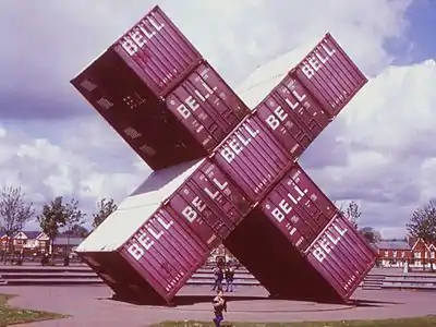 large-scale arrangement of red cuboid shipping containers in the shape of a letter X on display in a park with trees in the background. People walking underneath.