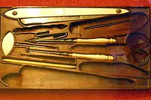 Photo of a very old waxed wood barber's kit with a dozen recessed compartments, in which some of the original tools are still to be found, including a razor, an awl and a needle. The scissors, knife and second razor are missing, thanks to the shapes in the wood. The blades are made of steel and the handles of horn or ivory.