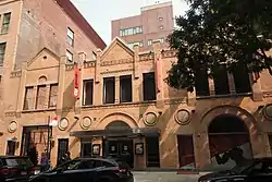 The right half of an old, sturdy-looking 4-story, 5-bay building, faced with partly soiled beige brick and architectural decorations. The right bay has a gabled fifth floor with 3 small windows.  A fire escape runs down the front. At street level 1½ large doorways are visible. One of the 20 or so windows is boarded, another is painted black.