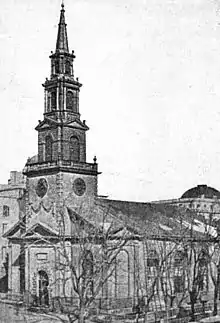 A black-and-white photograph of a church with a tall steeple, seen from its left across the street