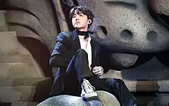 J-Hope sits on the foot of a stone lion. He is looking to the upper right, wearing a black suit and high-top converse.