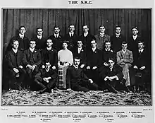 Group photograph of a seated woman in Victorian dress surrounded by 19 suited men.