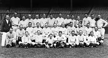 A posed team photo of a baseball team. They wear uniforms and are seated and standing in three rows, with a man in a jacket standing to the left.