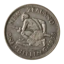 A coin featuring a crouched Māori atop a rocky escarpment. He holds a taiaha war-staff with both hands pointing it and facing forward off the escarpment. He is muscular and shirtless, but wears a piupiu skirt.