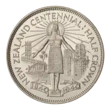 A coin featuring a Māori woman in a skirt standing between traditional and modern architecture. A sun and rays are behind her head. Below her is a ribbon reading "1840 – 1940". Around her is the encircling text "NEW ZEALAND CENTENNIAL HALF CROWN"