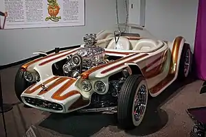 A white car with brown stripes, with open wheels and a clear bubble canopy over twin seats, and exposed, chromed engine with a blower. Beatnik Bandit II and a few of Roth's other cars are also on display in this museum.