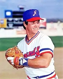 A man in a white baseball uniform with "Sounds" in blue and red across the front of the jersey and a blue cap with a white "N" on the center smiles with his hands together in his glove as if preparing to throw a ball.