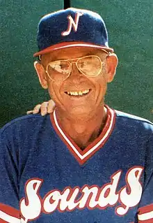 A man wearing glasses in a blue baseball jersey with "Sounds" written on the front in red and white smiles.