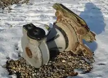 A large, dull-coloured, rust-flecked metal object lying on its side and with some broken wood attached to its base. It lies on a snow-covered beach on a patch of uncovered shingle.