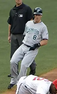 A man in a red baseball jersey with "Sounds" written on the front in white and blue and a blue cap with a white "N" on the center stands on a baseball field swinging a bat.