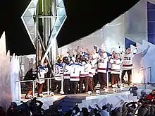 A group of men wearing white hockey jerseys surround a large torch. They light the torch and raise their arms in celebration.