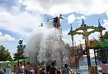 Line of people standing behind a fence looking at a colorful amusement park ride which is splashed by water.