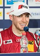 A man in his late twenties sporting a white baseball cap with a brand logo and dark red racing overalls and is speaking into a blue-coloured microphone at a press conference