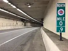 A two-lane highway running through a tunnel.  A green mile marker sign in the foreground reads NORTH / MILE 0.9