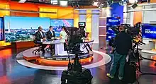 A look behind the scenes at a news set. Several people are seated at the desk. Two large cameras are visible, with a director standing between them.