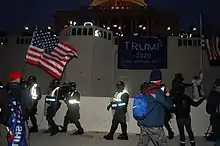 A photo taken near dusk of the Capitol building. A large Trump flag is hung on a railing. Protestors are still remain, and a small line of police officers yield shields.