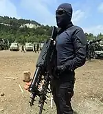 An armed militant of the Radwan Force. The militant is dressed in black tactical clothes and wears a black balaclava as well as dark goggles.