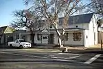 Single storey, 5-bay saddle roof house (now shop) with covered end gables and covered front gable with barge fret and 6 × 6 sash with four pilasters visible, c 1850 (F & C). Rectangular roof vents with plaster surrounds. Double door with small-pane Type of site: House Current use: House.