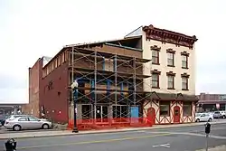 Two attached buildings, with a large parking lot around and behind them and an elevated highway in the background. The one on the right is brick, three stories high, painted yellow with an ornate decoration at the flat roof. On the left is another brick building with a sloping roof and scaffolding in the front.