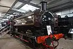 A red pannier tank locomotive, viewed from the front on display inside a railway museum, surrounded by other locomotives.