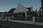 L-shaped, thatched house with mock-chimney end gables. The main gable faces the side and ia Worcester type with four pilasters and 12-pane casement, dated 1841. The street facade has a convex verandah on cast iron drip fret. 2 × 2 double door with upper, Type of site: House Current use: House.