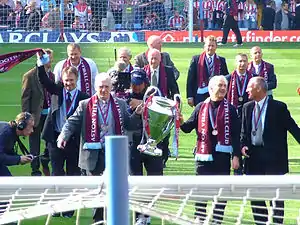 In the foreground is two men holding a large cup, they have claret scarves and a medal around their necks. Around them are ten old players in suits with medals and scarves around their necks