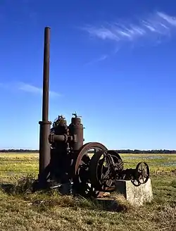 Photo of a very rusted industrial-looking pump with tall tube and wheel sits in a flat rice field.