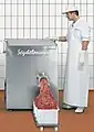 A man stands before a chest-high boxy machine that is pushing meat into an overflowing plastic bin on wheels.