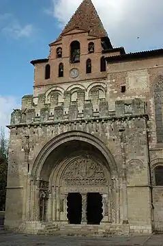 A very large porch of yellowish stone, with a single enormous, slightly pointed archway, juts from the side of a building.