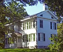 A white house with green shutters and gray roof in the middle of some trees