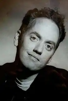 A black-and-white image of a man with short dark hair and a sweater with a white shirt underneath. He is looking toward the camera.