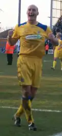 A bald man wearing yellow football shirt, shorts and socks and black football boots. He is smiling broadly and clenching his fists. He is standing on a grass surface and several people and indistinct structures are behind him.