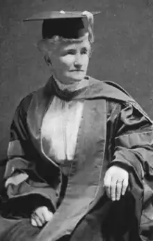 An old white woman, seated, wearing academic robes and a mortar board cap