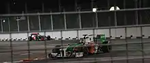 a white, green, and orange F1 car drives down along a closed street at night ahead of a silver F1 car