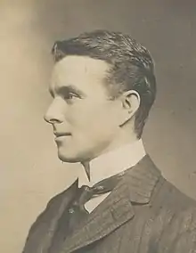 A man, fresh-faced with dark, brushed-back hair, seated among a group. He is wearing a naval officer's uniform with a high, stiff collar