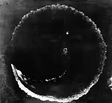 Photograph looking directly down at the ocean's surface from a high altitude. There is a rough circle on the surface that is about as wide as the photograph. The top deck of a ship is seen at the lower left of the circle; the length of the ship is much smaller than the circumference of the circle.