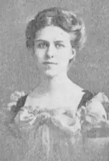 A young white woman with hair parted center and dressed in an updo, wearing a dress with a square neckline and elaborate draping