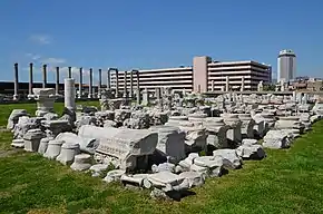 Rectangular stone ruin, with damaged columnade behind alongside modern building.