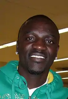 A black man wearing a green hooded top and white shirt smiles into a camera.