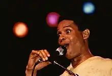 A man with his eyes wide open, holding a microphone placed on a stand. In the background are three stage lights, each a different color