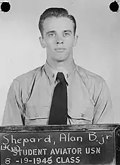 Shepard, in Navy uniform short and tie, stands before a blackboard on which is stencilled "Student aviator USN – 8-19-1946 class. Above that is written in chalk: "Lt (jg) Shepard, Alan B. Jr"