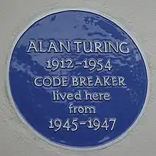 A Blue plaque on a white wall with the words "Alan Turing 1912–1954 CODE BREAKER lived here from 1945 – 1947