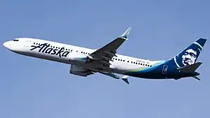 A white plane with a navy blue eskimo on the tail and the word "Alaska" painted across the fuselage, in-flight over a blue sky