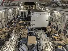 Photograph of the inside of a cargo jet, containing people in military field uniforms, equipment, and trucks