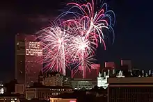 Blue and red fireworks explode over a complex of buildings after dusk.