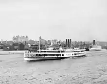 A white steam ship is seen near the shore of the Hudson River in front of the downtown area of Albany; the New York State Capitol can be seen in the background.