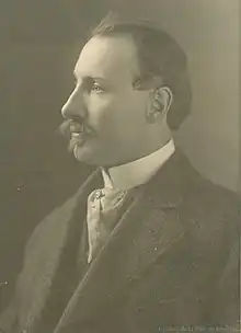 A sepia image of a white man with dark hair, an bushy moustache and goatee. In a heavy suit jacket and wearing a cravat, he looks to the distance, off to the side, showing his left cheek.