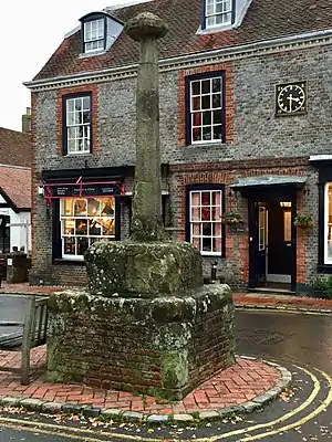 The cross is formed of stone base and shaft on a brick plinth; the shaft is topped with a ‘cornice-like’ stone