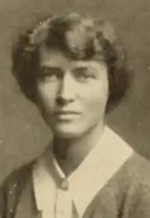 A young white woman with short wavy hair, wearing a white blouse with a wide pointed collar, and a soft jacket or cardigan